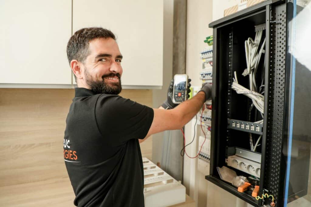 Électricien Luc Energies souriant utilisant un multimètre sur un tableau électrique pour une installation sécurisée.
